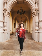 Fototapeta na wymiar Brunette girl in a red jacket and with a backpack stands near the arch with beautiful stucco and architecture in the Park in San Diego