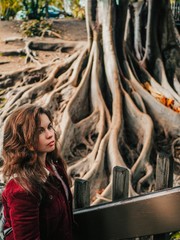 Girl with long hair in a red jacket stands against the background of huge trees with giant roots on the surface in San Diego