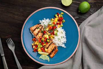 Grilled salmon steak with fruit sauce and rice on a blue plate. Delicious meal made of fish and exotic fruits and vegetables on a table. Top view shot above.