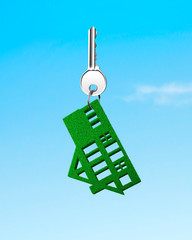 Buying green energy house concept. Silver key with green grass house shape keyring, on blue sky background.
