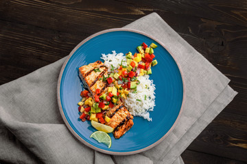 Grilled salmon fillet with fruit sauce made of mango, bell pepper, onion and avocado with basmati rice garnish on a blue plate. Delicious seafood meal. Top view shot above.
