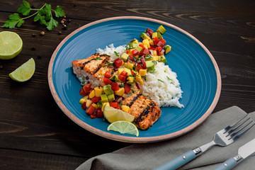 Salmon fillet cooked on grill with exotic sauce made of mango, avocado and pepper on blue plate. Delicious fish meal with jasmine rice on a table.