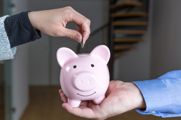 Frau steckt eine Münze in den Sparschwein, den ein Mann in seiner Hand festhält, sie sparen für eine Eigentumswohnung