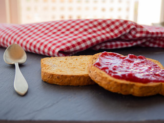 Two slices of toast with raspberry jam on a slate plate, a spoon and a tablecloth old style