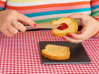 A woman with painted nails smearing red raspberry jam with a knife on a slice of toast