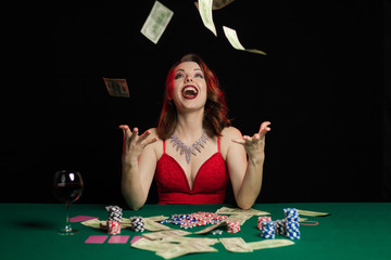 Emotional young lady in an evening red dress playing cards on a table on green cloth in a casino