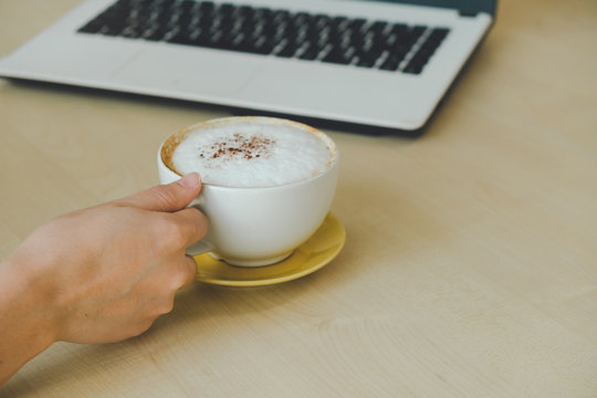 Closeup image of woman hand holding a cup of hot coffee on wooden table, working with computer laptop, freelance and work concept