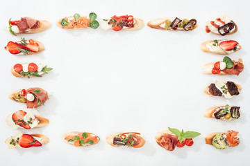 top view of frame made of traditional italian bruschetta with prosciutto, salmon, fruits, vegetables and herbs on white with copy space