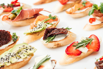 selective focus of italian bruschetta with dried tomatoes, prosciutto, avocado, strawberries and herbs on white