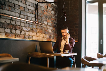 Young handsome man sitting in office with cup of coffee and working on project connected with modern cyber technologies. Businessman with notebook trying to keep deadline in digital marketing sphere.
