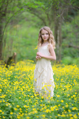 A cute young girl is in the forest on the edge of the forest with yellow flowers. She has blond curly hair, she's wearing a beautiful dress.