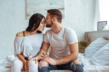 handsome man in jeans and beautiful woman sitting on bed and hugging