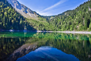 Fototapeta na wymiar Symmetrical reflections. Turquoise water of a mountain lake surrounded by green wooded hills under a blue sky. Lake Ritsa Tourism in the Caucasus in Abkhazia.