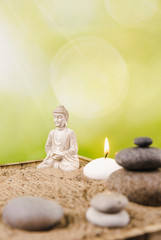 Miniature desk zen sandbox with Buddha figure sit in Lotus position, stacked zen sea stones, spa candles burning against green bokeh studio background, copy space.