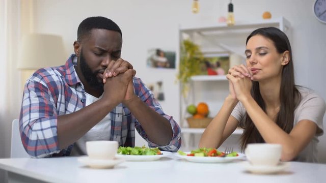 Young mixed-race couple praying before lunch, vegetarian healthy salad on table