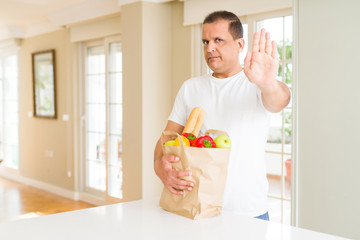 Middle age man holding groceries shopping bag at home with open hand doing stop sign with serious and confident expression, defense gesture