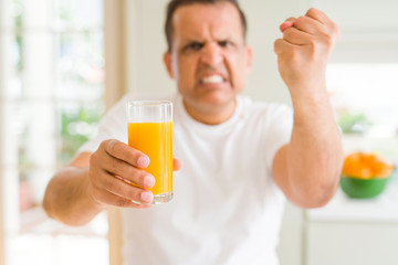 Middle age man drinking a glass of orange juice at home annoyed and frustrated shouting with anger, crazy and yelling with raised hand, anger concept