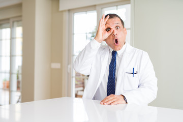 Fototapeta premium Middle age doctor man wearing medical coat at the clinic doing ok gesture shocked with surprised face, eye looking through fingers. Unbelieving expression.