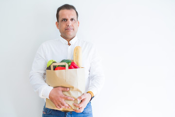 Middle age man holding groceries shopping bag over white background with a confident expression on smart face thinking serious