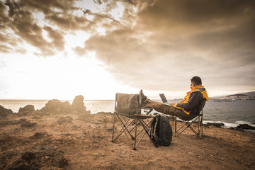 Travel and enjoying outdoor people concept with lonely man working on laptop internet connecetd computer sitting in front of an amazing sunset on the ocean - digital nomad millennial concept