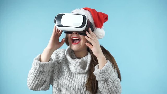 Woman wearing santa claus hat exploring space with virtual reality goggles headset. Amazed girl watching 3d film tour in vr glasses box. New generation cyber christmas concept, on blue