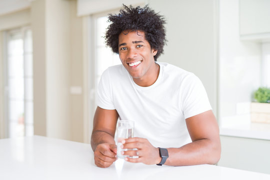 African American man drinking a glass of water at home with a happy face standing and smiling with a confident smile showing teeth
