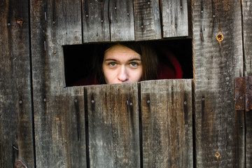 Young woman locked in a wooden old barn, sad looks out the slit.