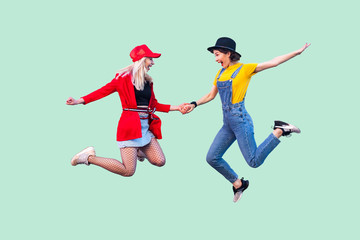 Fototapeta na wymiar Full legs body size portrait of two happy screaming stylish hipster girls in fashionable clothes are jumping up in the air and celebrated a win. Indoor studio shot, isolated on green background