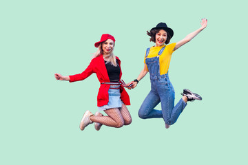 Full legs body size portrait of two happy screaming stylish hipster girls in fashionable clothes are jumping up in the air and looking at camera. Indoor studio shot, isolated on green background