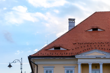 Traditional roof with small windows like eyes in Sibiu, Transylvania, Romania. Detailed architecture of a house top with columns, chimney, and a street pole, on pale light blue sky with white clouds.