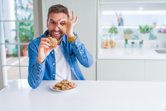 Handsome man eating chocolate chips cookies with happy face smiling doing ok sign with hand on eye looking through fingers