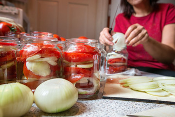 Canning fresh tomatoes with onions in jelly marinade. Woman hands putting red ripe tomato slices and onion rings in jars. Basil, parsley leaves on top of onions. Vegetable salads for winter