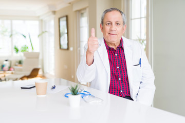 Handsome senior doctor man wearing medical coat at the clinic doing happy thumbs up gesture with hand. Approving expression looking at the camera with showing success.