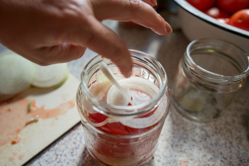 Canning fresh tomatoes with onions in jelly marinade. Woman hands putting red ripe tomato slices and onion rings in jars. Basil, parsley leaves on top of onions. Vegetable salads for winter