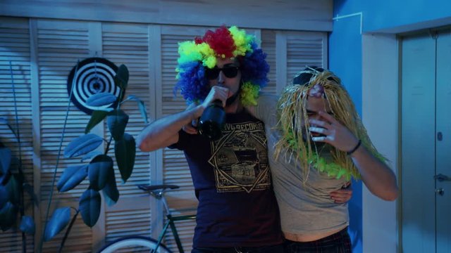 Two funny students coming home after&nbsp;celebrating Birthday. Happy young men in colored clown wig and female blonde wig. Night, indoors, courtesy, concept, heavy drinking