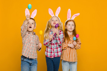 surprised two little girls and boy with Easter bunny ears holding colorful eggs on yellow background. Happy easter. cute children