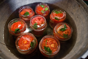Canning fresh tomatoes with onions in jelly marinade. Conservation and cooking. Jars boiling in water to kill bacteria. Sealing process.  Basil, parsley leaves on top. Vegetable salads for winter