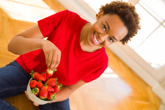 Beautiful young african woman with afro hair eating fresh strawberries sitting on the floor