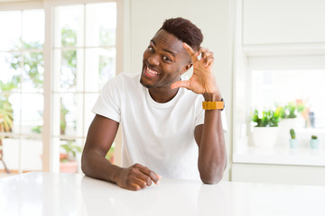 Handsome african american man on white table at home smiling and confident gesturing with hand doing size sign with fingers while looking and the camera. Measure concept.