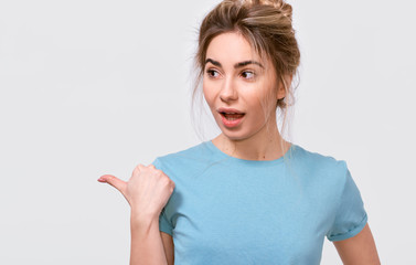European excited young woman wearing casual blue t-shirt, indicating to blank copy space for advertising text, looking aside, posing over gray studio background. People emotions