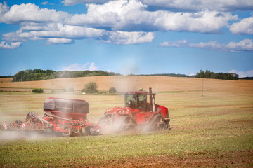 Fototapeta premium Agricultural background with red tractor pulling plow, throwing dust in air. Combine harvester at wheat field. Heavy machinery during cultivation, working on fields. Dramatic sky, rain, storm clouds 