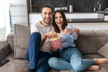 Image of excited couple eating popcorn from bucket while sitting on couch indoor and watching movie