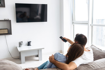 Image from back of attractive couple in casual wear watching television on sofa at home