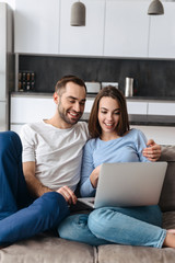 Image of satisfied couple using laptop together while sitting on sofa in living room at home