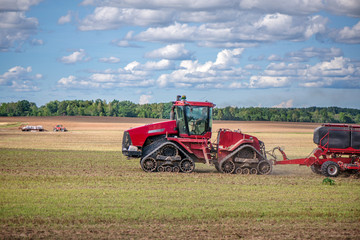 Agricultural background with red tractor pulling plow, throwing dust in air. Combine harvester at wheat field. Heavy machinery during cultivation, working on fields. Dramatic sky, rain, storm clouds
