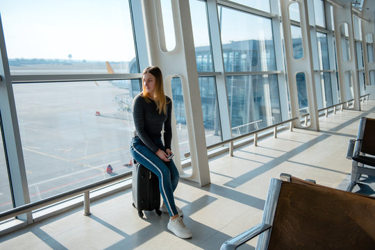 Young female passenger sitting on luggage bag in the airport terminal hall while waiting her flight. Woman near the big windows
