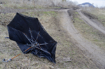 An old, rusty, broken black umbrella lies on the side of a country road.