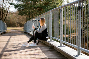 Young woman in casual clothes sitting with a bicycle on the wooden bridge near the lake in the countryside on sunny day and drinking water