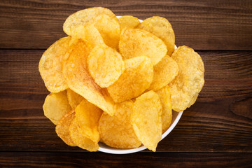 Crispy potato chips in bowl on wooden background, top view