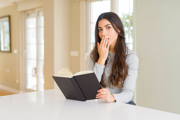 Young woman reading a book cover mouth with hand shocked with shame for mistake, expression of fear, scared in silence, secret concept